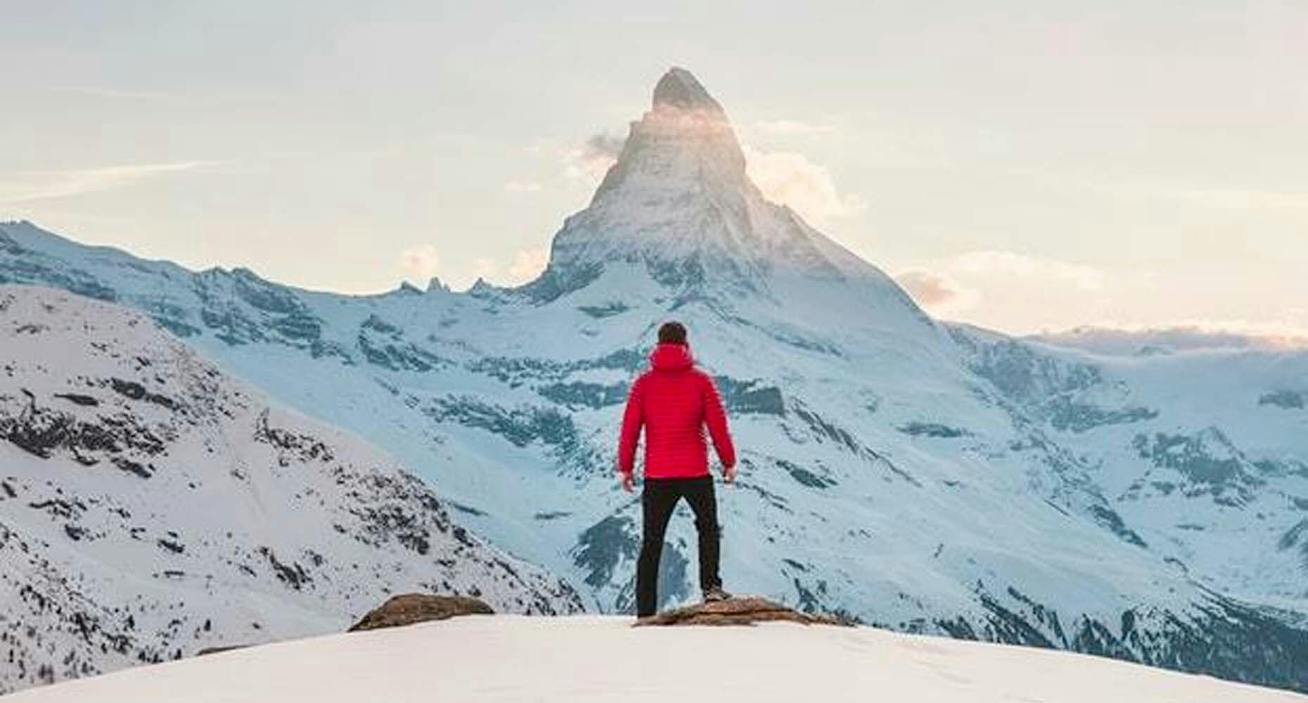 Man in red on a mountain looking at a bigger mountain